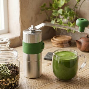 May include: A cylindrical manual coffee or tea grinder made of brushed silver metal with a green silicone grip band and a matching green handle knob. The grinder features a long metal crank arm positioned above a clear glass mug filled with green tea.