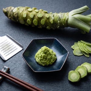 May include: A close-up shot of fresh wasabi root, grated wasabi, and sliced wasabi. The wasabi is green and sits on a dark blue hexagonal plate. A metal grater and chopsticks are also in the image.