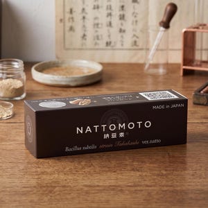 May include: A rectangular dark brown box labelled NATTOMOTO with white text including Bacillus subtilis strain Takahashi ver.natto and Made in Japan. The packaging features a circular logo, a QR code, and small circular graphic icons on the top surface.