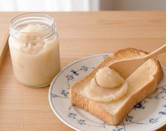 May include: A jar of light-colored spread next to a slice of toasted bread with a wooden spoon. The bread is on a white plate with a blue floral pattern. The jar is clear glass.