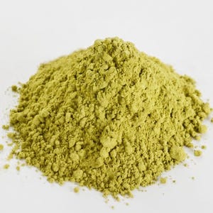 Pode incluir: Um monte de pó fino de cor verde-oliva claro com uma textura seca e granulada. A substância está empilhada em forma cónica, exibindo uma cor terrosa consistente em todo o monte, com pequenos grumos visíveis por toda a superfície.