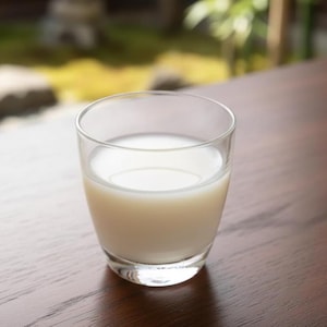 May include: A transparent glass containing a milky white liquid, possibly a drink. The glass rests on a dark wooden table, with a blurred outdoor scene in the background.