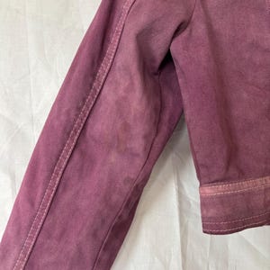May include: Close-up of a dusty rose denim jacket sleeve. The sleeve has white stitching along the seams and cuffs. The fabric shows signs of wear and discoloration, adding to its vintage aesthetic. The jacket is made of a sturdy denim material.