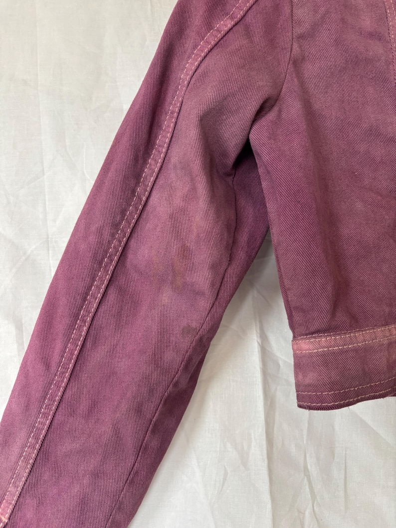 May include: Close-up of a dusty rose denim jacket sleeve. The sleeve has white stitching along the seams and cuffs. The fabric shows signs of wear and discoloration, adding to its vintage aesthetic. The jacket is made of a sturdy denim material.