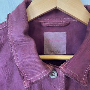 May include: Close-up of a dusty rose denim jacket with a classic collar and two silver buttons. The jacket has a distressed look with visible stitching and a label that reads "Free People". The jacket is hanging on a wooden hanger.