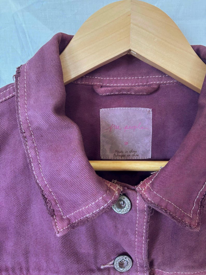 May include: Close-up of a dusty rose denim jacket with a classic collar and two silver buttons. The jacket has a distressed look with visible stitching and a label that reads "Free People". The jacket is hanging on a wooden hanger.