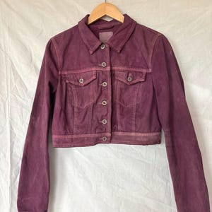 May include: A cropped, burgundy denim jacket with a classic collar, button-down front, and two chest pockets. The jacket is hanging on a wooden hanger against a white background. The fabric has a slightly faded appearance.