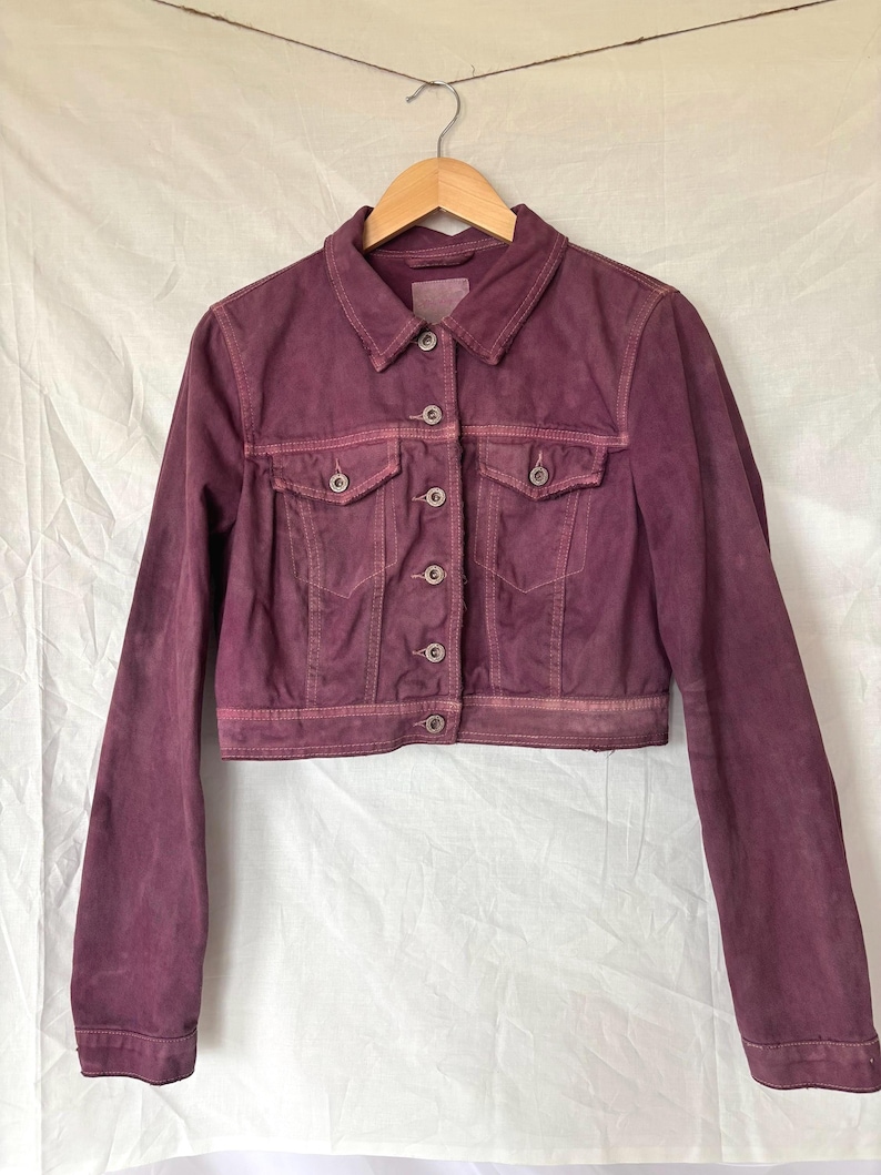 May include: A cropped, burgundy denim jacket with a classic collar, button-down front, and two chest pockets. The jacket is hanging on a wooden hanger against a white background. The fabric has a slightly faded appearance.