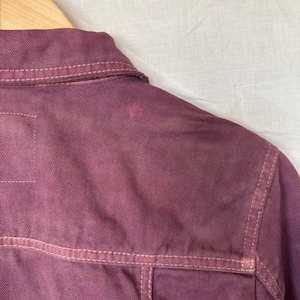 May include: A close-up view of a burgundy denim jacket, focusing on the back and shoulder area. The jacket has lighter-coloured stitching and a small stain. It is suspended on a wooden hanger.