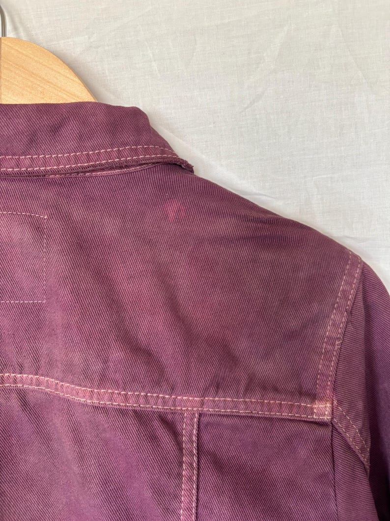 May include: A close-up view of a burgundy denim jacket, focusing on the back and shoulder area. The jacket has lighter-coloured stitching and a small stain. It is suspended on a wooden hanger.