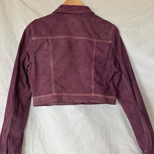 May include: A cropped, burgundy denim jacket with a classic collar and button fastenings. The jacket has a faded, distressed appearance with visible stitching details. It is hanging on a wooden hanger against a white backdrop.