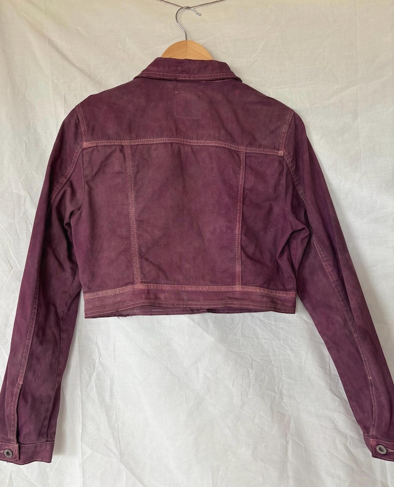May include: A cropped, burgundy denim jacket with a classic collar and button fastenings. The jacket has a faded, distressed appearance with visible stitching details. It is hanging on a wooden hanger against a white backdrop.