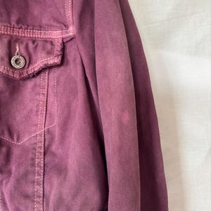 May include: Close-up of a dusty rose denim jacket. The jacket has a button closure, a chest pocket, and visible stitching. The fabric has a slightly faded appearance, with a few lighter spots on the sleeve.