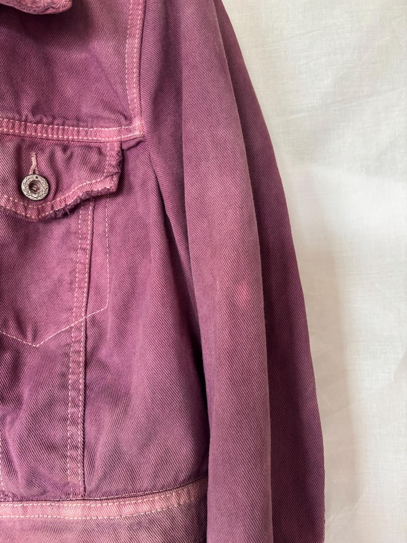 May include: Close-up of a dusty rose denim jacket. The jacket has a button closure, a chest pocket, and visible stitching. The fabric has a slightly faded appearance, with a few lighter spots on the sleeve.