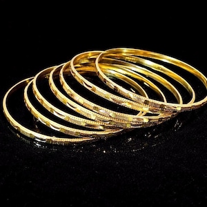 May include: A collection of gold bangles with intricate etched designs. The bangles are stacked together, reflecting light against a black background. The jewelry is made of a shiny, yellow metal.