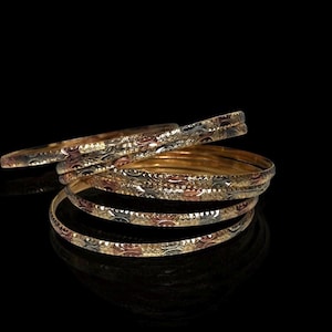 Könnte beinhalten: Eine Sammlung goldener Armreifen mit aufwendigen Mustern. Die Armreifen sind auf einer reflektierenden schwarzen Oberfläche gestapelt und angeordnet. Die Armreifen haben eine Mischung aus Gold-, Silber- und Roségoldtönen.