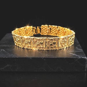 May include: A gold bracelet featuring a series of interconnected rectangular links with a textured, nugget-like surface. The bracelet has a polished finish that reflects light, creating a bright, metallic appearance across the repeating segments.