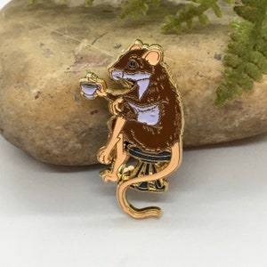 May include: A gold-colored enamel pin featuring a brown rat seated on a small stool, holding a teacup. The rat is wearing a brown coat with a light purple vest. The pin has a detailed design and is set against a neutral background.