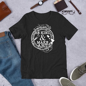 May include: Dark gray t-shirt with the text "BE KIND & MAKE ART" in a white circular design. The design includes a paintbrush, paint tube, and other art supplies. The shirt is displayed on a flat surface with jeans and accessories.