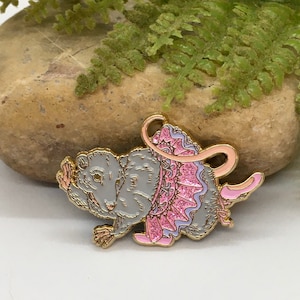 Könnte beinhalten: Ein grauer Ratten-Emaille-Pin mit goldenen Verzierungen, der einen rosa Tüllrock mit Glitzer und eine rosa Schleife trägt. Die Ratte befindet sich in einer spielerischen Pose, mit erhobener Vorderpfote. Der Pin ist vor einem neutralen Hintergrund platziert.