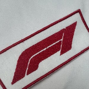 Könnte beinhalten: Ein rechteckiger, gestickter Aufnäher mit dem roten Formel-1-Logo auf weißem Hintergrund. Der Aufnäher hat einen dicken, roten gestickten Rand und ist auf eine weiße Stoffoberfläche mit einer sichtbaren Webstruktur genäht.