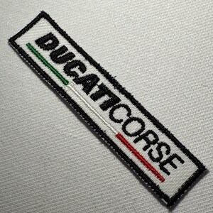 Op de afbeelding: Een rechthoekige geborduurde patch met de zwarte tekst DUCATICORSE op een witte achtergrond. Het ontwerp bevat een dunne horizontale lijn onder de tekst, gekleurd in groen, wit en rood, allemaal omsloten door een dikke zwarte rand.