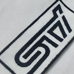 Peut inclure: Un écusson rectangulaire brodé présentant les lettres STI dans une police noire, grasse et stylisée sur un fond blanc. L'écusson possède une bordure noire épaisse et est fixé sur un tissu gris clair à la texture tissée visible.