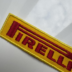 Może przedstawiać: Prostokątna, żółta naszywka z haftowanym napisem "PIRELLI" w kolorze czerwonym. Naszywka ma żółtą obwódkę i znajduje się na białej powierzchni.