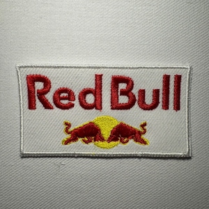 Può includere: Una toppa rettangolare ricamata su uno sfondo di tessuto bianco. La toppa presenta la scritta rossa Red Bull sopra un grafico di un sole giallo con due tori rossi che si fronteggiano in posizione di carica, il tutto racchiuso da un bordo cucito.