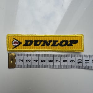 Könnte beinhalten: Ein rechteckiger, gelber Stickaufnäher mit dem schwarzen Schriftzug DUNLOP und einem Logo, das einen roten Buchstaben D in einem weißen Kreis zeigt. Ein unter dem Aufnäher platziertes Maßband zeigt eine Länge von ca. 10,8 cm.