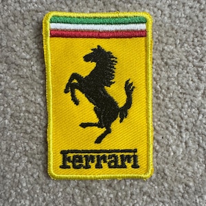Może przedstawiać: Żółta prostokątna naszywka z logo Ferrari. Zawiera czarnego, wspinającego się konia i napis "Ferrari" w czarnych literach. Górna krawędź ma zielone, białe i czerwone paski.