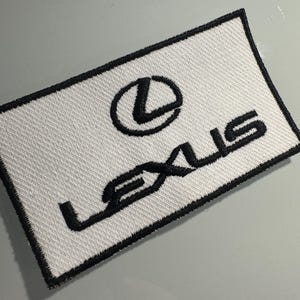 Może przedstawiać: Prostokątna haftowana naszywka z białym tłem i czarną obwódką. Wzór zawiera czarne logo Lexus oraz słowo LEXUS napisane czarnymi wielkimi literami, wyśrodkowane na powierzchni tkaniny z wyraźną teksturą ściegu.