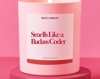 May include: A lit pink soy wax candle in a matching container, labeled "Smells Like a Badass Coder" with "Maiye Candles" at the top. The candle sits on a pink pedestal, with a pink background. The candle is 9 oz.