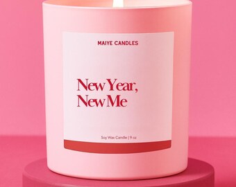 May include: A lit pink candle in a matching container sits on a round pedestal. The candle has a white label with the text "MAIYE CANDLES" and "New Year, New Me." The label also reads "Soy Wax Candle | 9 oz."