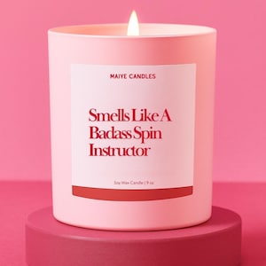 May include: A lit pink soy wax candle in a matching container, labeled "Smells Like A Badass Spin Instructor." The candle, from Maiye Candles, is 9 oz and sits on a pink pedestal against a pink backdrop.