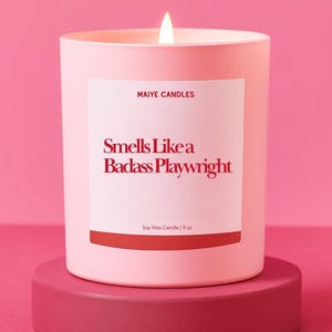 May include: A light pink soy wax candle with the text "Smells Like a Badass Playwright" on a white label. The candle is 9 oz and sits on a matching pink pedestal, with a lit wick.