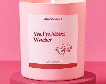 May include: A lit pink soy wax candle with the text "Yes, I'm A Bird Watcher" and a graphic of binoculars. The candle is 9 oz and sits on a matching pink pedestal, against a pink background.
