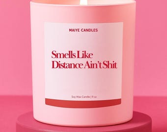 May include: A light pink soy wax candle in a cylindrical glass jar, with a white label that reads "Smells Like Distance Ain't Shit" in red. The candle is 9 oz and sits on a matching pink pedestal against a pink background.