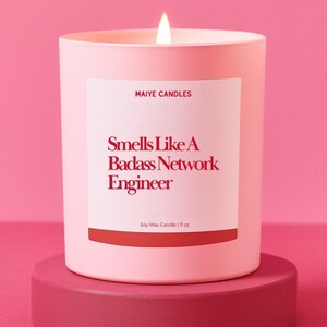 May include: A lit pink candle in a matching container, labeled "Smells Like A Badass Network Engineer." The candle sits on a pink pedestal against a pink backdrop. The label also reads "Maiye Candles" and "Soy Wax Candle | 9 oz."