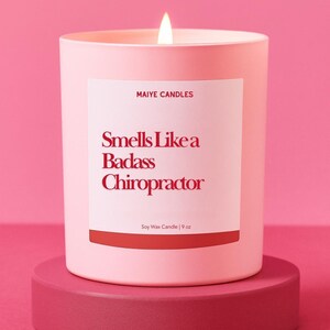 May include: A light pink soy wax candle in a matching pink container, with a lit wick. The label reads "Smells Like a Badass Chiropractor" and "Maiye Candles". The candle is 9 oz and sits on a pink pedestal.