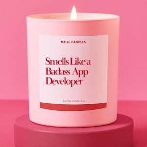 May include: A lit pink soy wax candle in a matching container, with the text "Smells Like a Badass App Developer" in red. The candle is 9 oz and sits on a pink pedestal against a pink background. The brand name "Maiye Candles" is printed above the text.