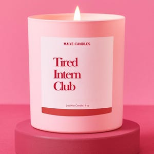 May include: A lit pink soy wax candle in a matching container, labeled "Tired Intern Club" in red text. The candle sits on a round, dark pink pedestal against a pink backdrop. The candle is 9 oz.