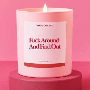 May include: A lit pink soy wax candle in a matching pink container, sitting on a pink pedestal. The candle has a white label with the text "Fuck Around And Find Out" in red, and "MAIYE CANDLES" above it. The candle is 9 oz.