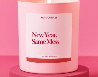 May include: A lit pink soy wax candle in a cylindrical container, labeled "New Year, Same Mess" with "Maiye Candles" above. The candle sits on a matching pink pedestal against a pink backdrop. The candle is 9 oz.