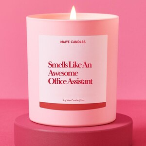 May include: A lit pink soy wax candle in a matching pink container. The label reads "Smells Like An Awesome Office Assistant" with the brand name "Maiye Candles" above. The candle is 9 oz and sits on a pink pedestal.