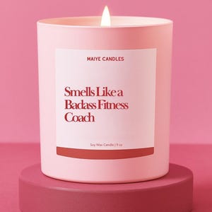 May include: A pink soy wax candle with the text "Smells Like a Badass Fitness Coach" on a white label. The candle is 9 oz and sits on a matching pink pedestal. The candle is lit with a bright flame.