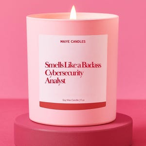 May include: A lit pink candle on a matching stand. The candle has a white label with the text "Smells Like a Badass Cybersecurity Analyst." The candle is 9 oz and made of soy wax. The brand name "Maiye Candles" is also on the label.