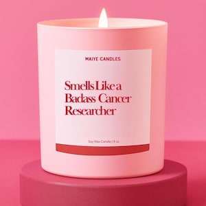 May include: A light pink candle with a lit wick, labeled "Smells Like a Badass Cancer Researcher." The candle is in a matching pink container and sits on a darker pink pedestal. The label also reads "Maiye Candles" and "Soy Wax Candle | 9 oz."