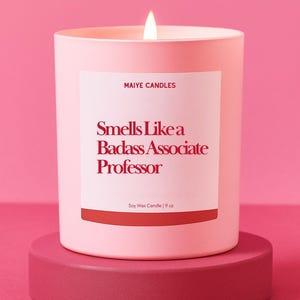 May include: A lit pink soy wax candle in a matching pink container, sitting on a pink pedestal. The label reads "Smells Like a Badass Associate Professor" and "9 oz". The candle is from Maiye Candles.