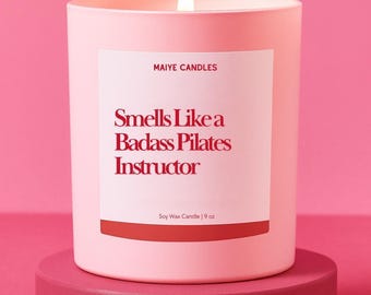 May include: A lit pink candle on a matching pink pedestal. The candle has a white label with the text "Smells Like a Badass Pilates Instructor" in red. The label also includes the text "MAIYE CANDLES" and "Soy Wax Candle | 9 oz."